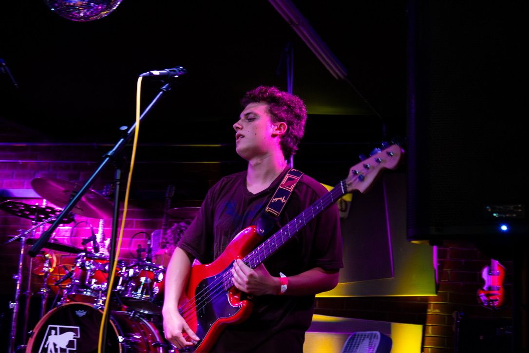 Jake Horvitz playing a red bass guitar on stage under magenta and amber lights, a drum kit behind him.